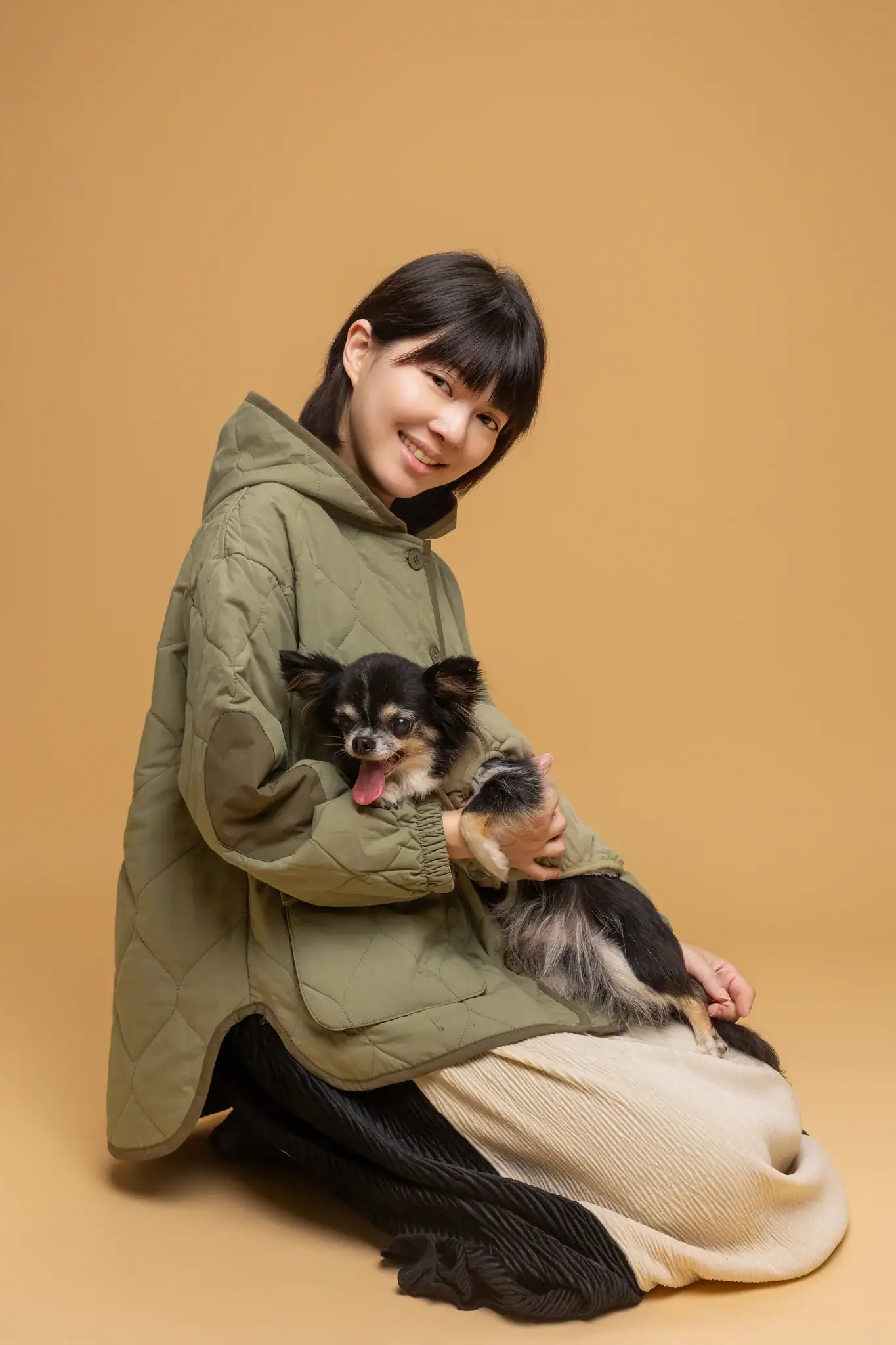 Lisa 身穿墨綠色外套，跪姿抱著黑棕吉娃娃，兩者在溫暖的米色背景前一同微笑面對鏡頭。