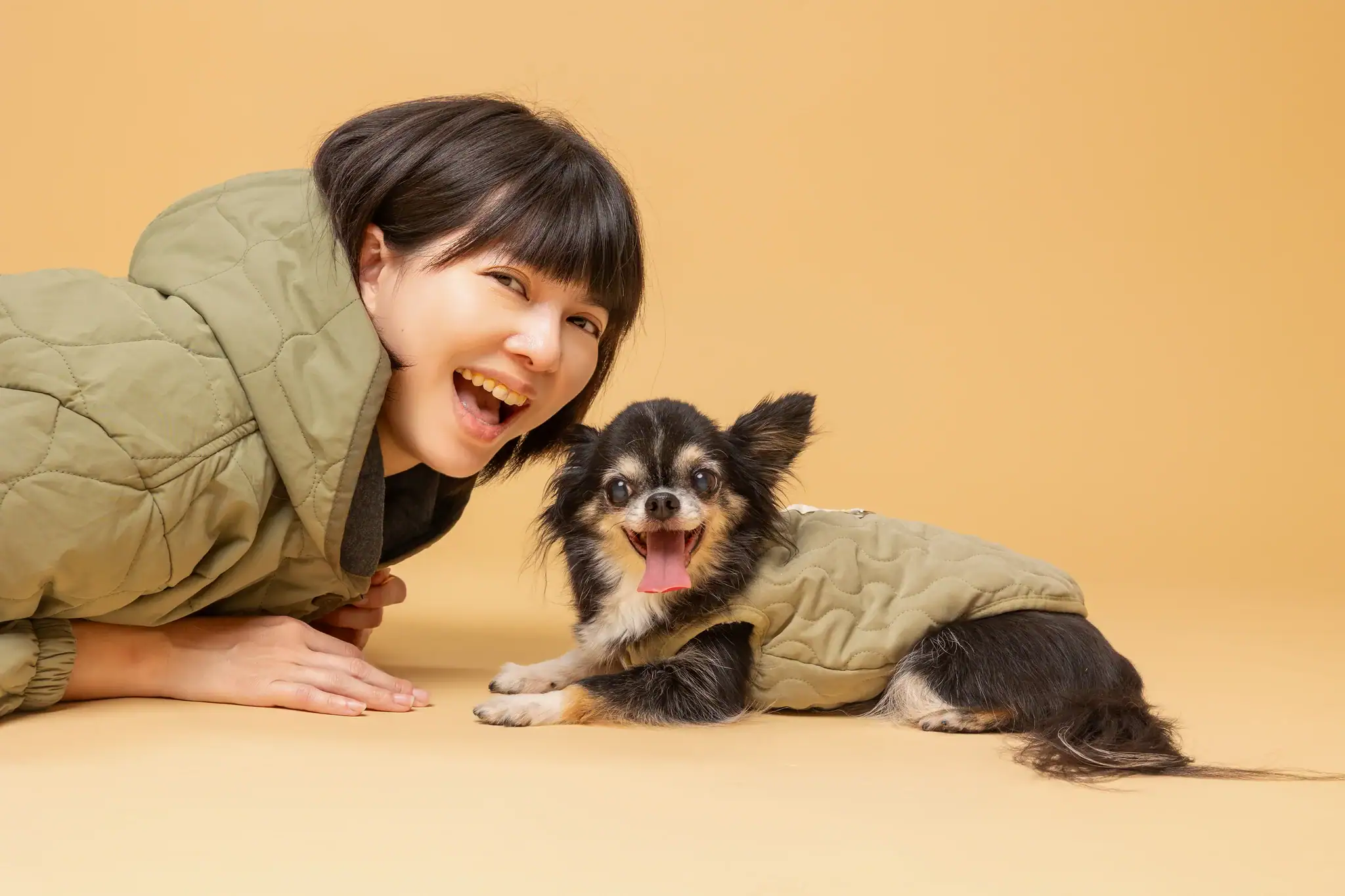 Lisa 趴臥在米色地板上，身旁是穿著綠色小外套的黑棕吉娃娃，兩人開心地大笑，充滿了歡樂氛圍。