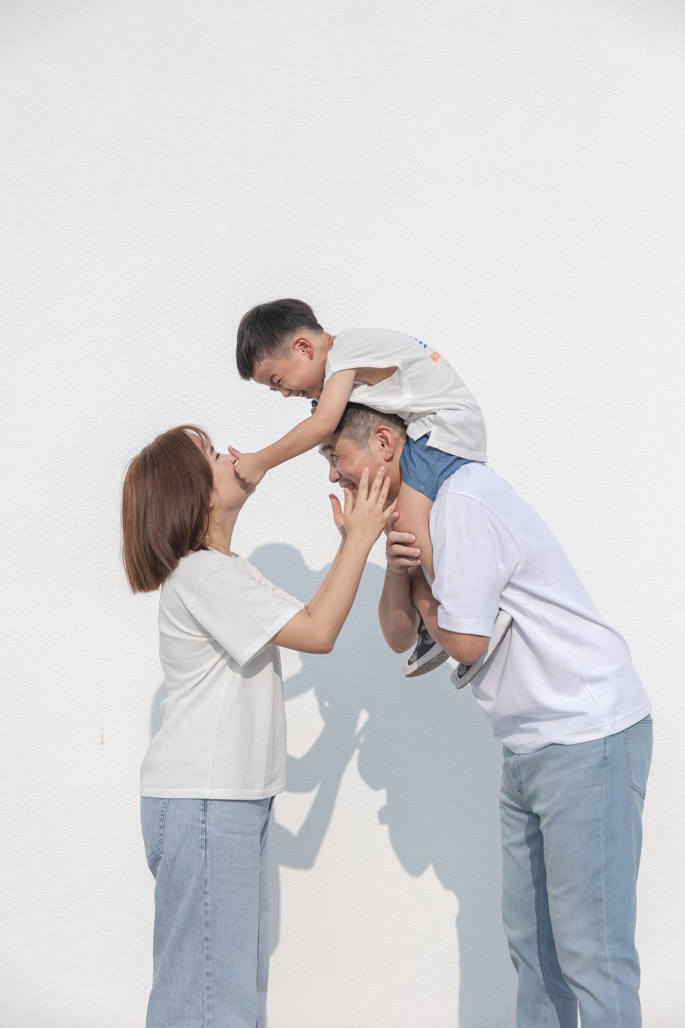 爸爸把品捷扛在肩上、媽媽在旁邊逗他笑的白牆前親子寫真