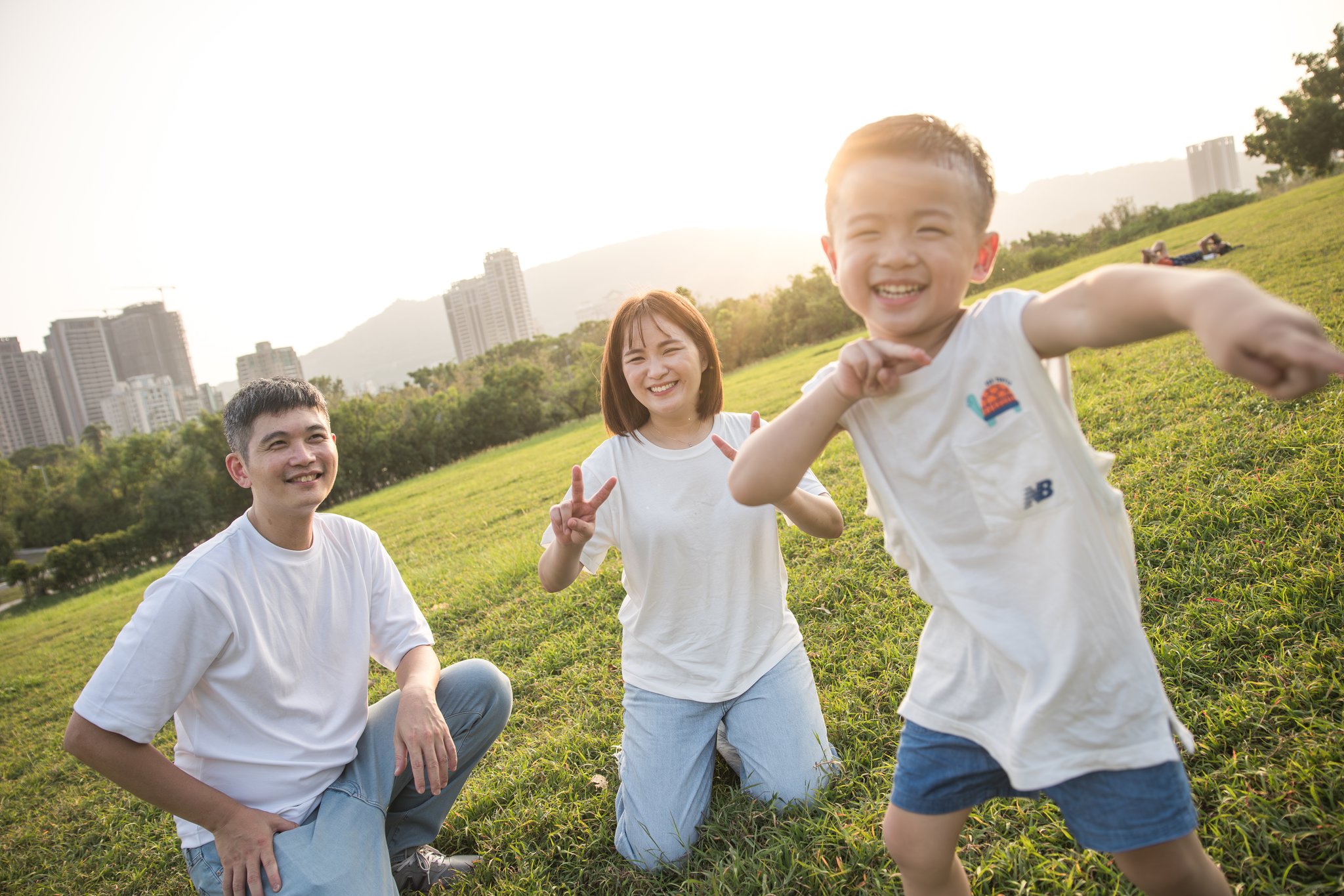 夕陽逆光中孩子張開雙手大笑，爸媽蹲在後方陪著
